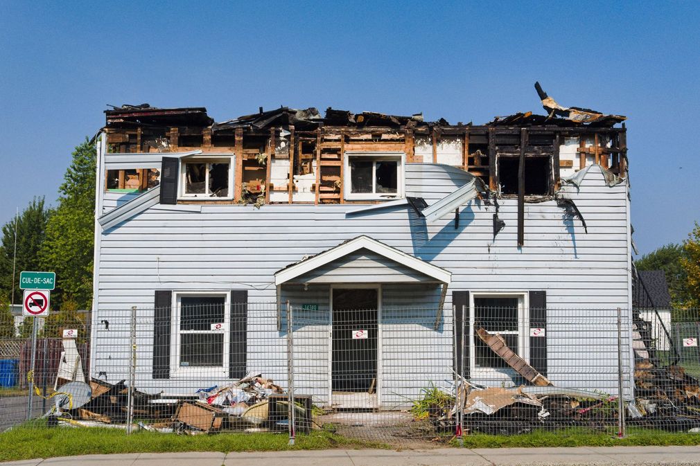 Burned two-story white house with roof and upper windows heavily damaged, standing behind a fence.