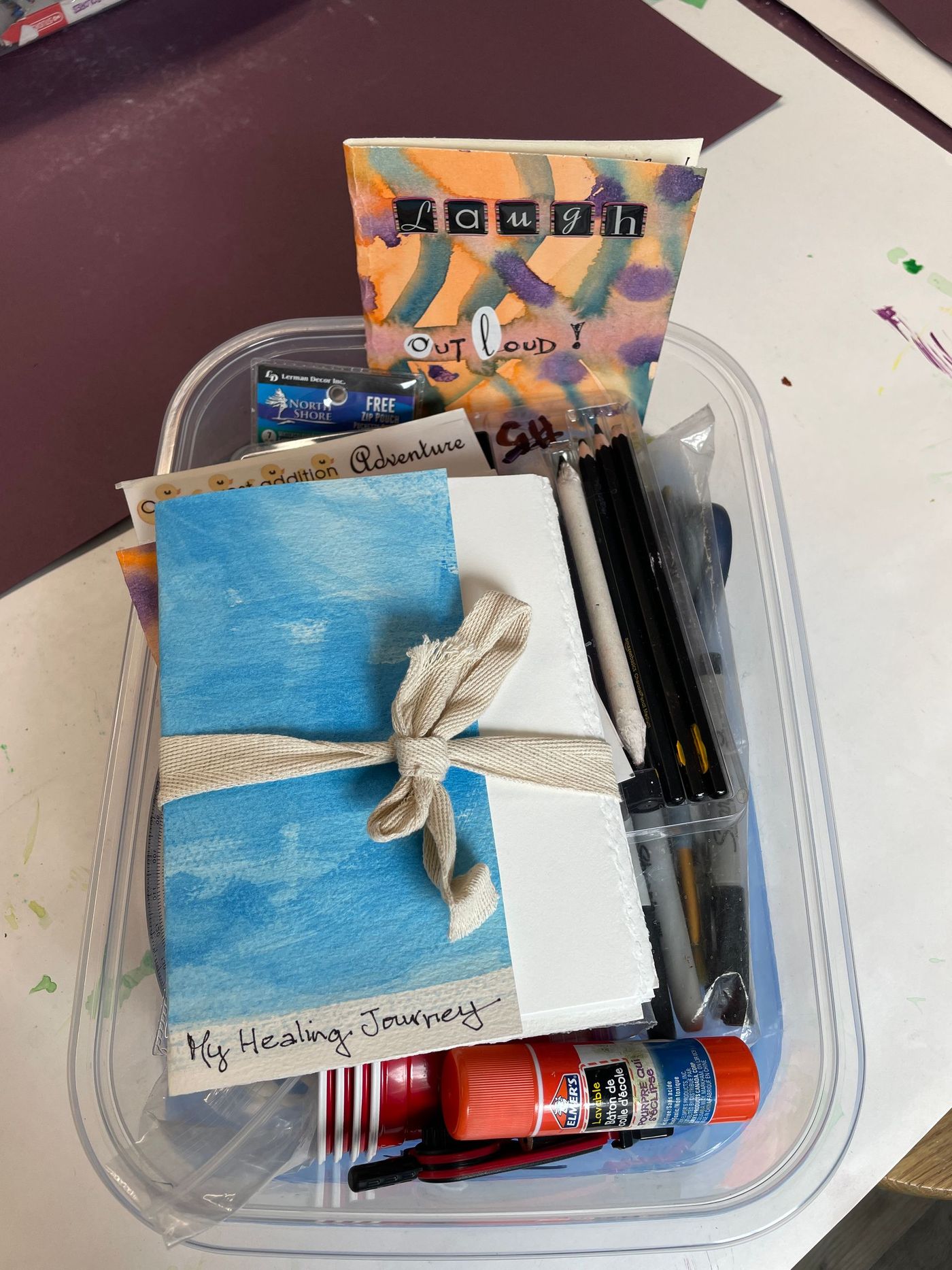 Clear plastic tray with assorted toiletries,  a blue notebook, rope tie, and black pens for grief journey at home art