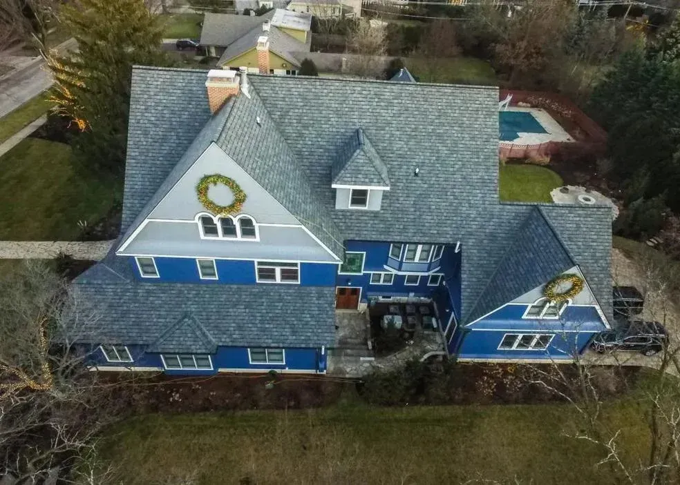 An aerial view of a large, blue, two-story house with a grey shingled roof and decorative holiday wreaths.
