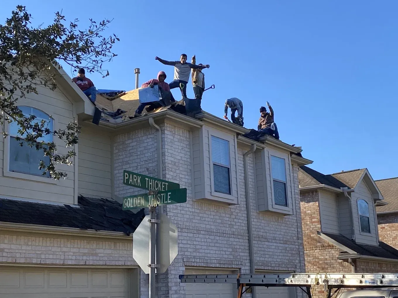 A group of workers on a suburban house roof, some waving or sitting, on a sunny day next to a street sign.