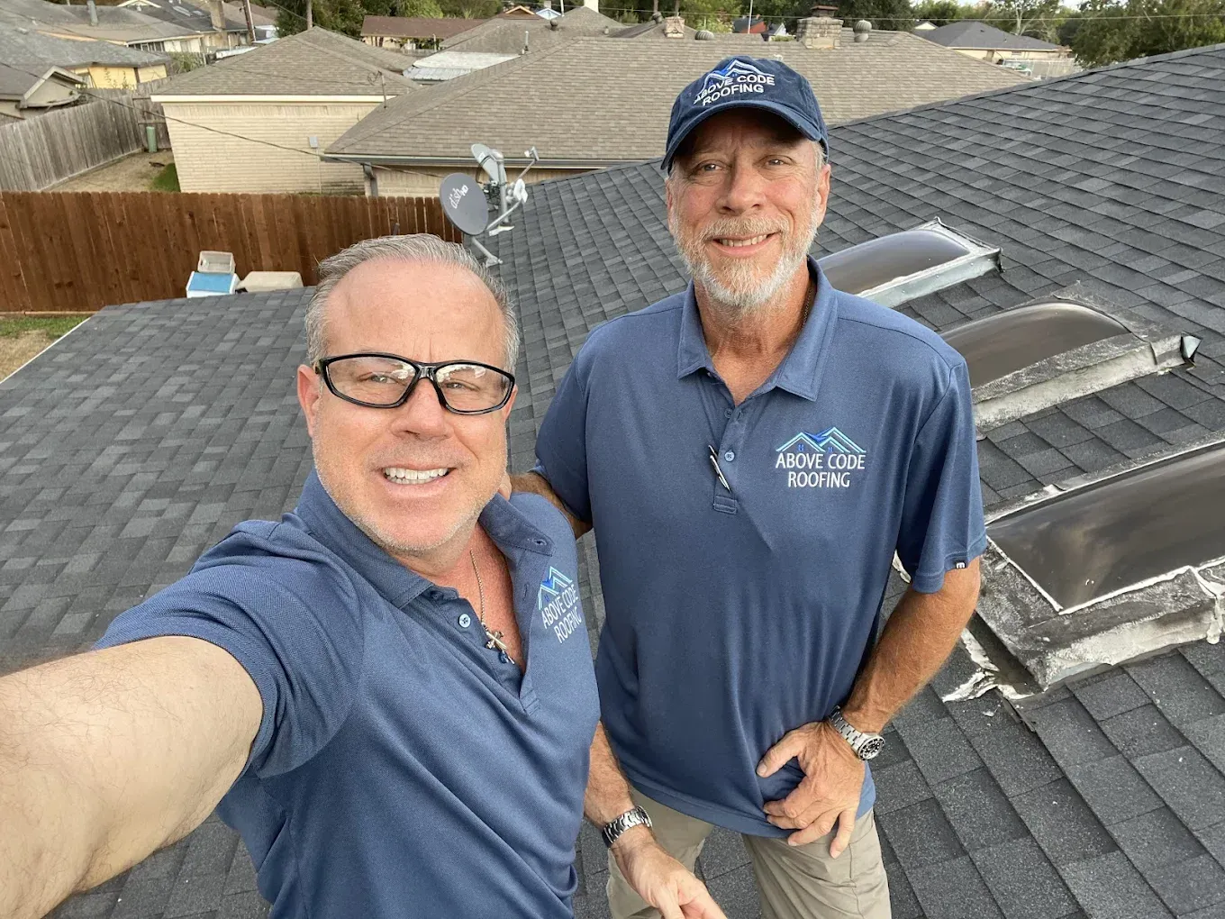 Two people in blue branded shirts stand on a dark shingled roof, one taking a selfie and the other smiling.