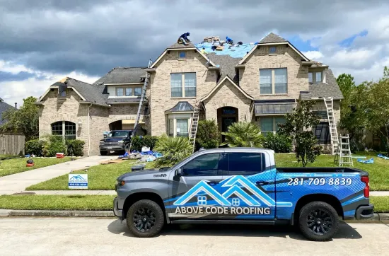 A grey Above Code Roofing truck parked in front of a multi-story brick house during a roofing repair project.