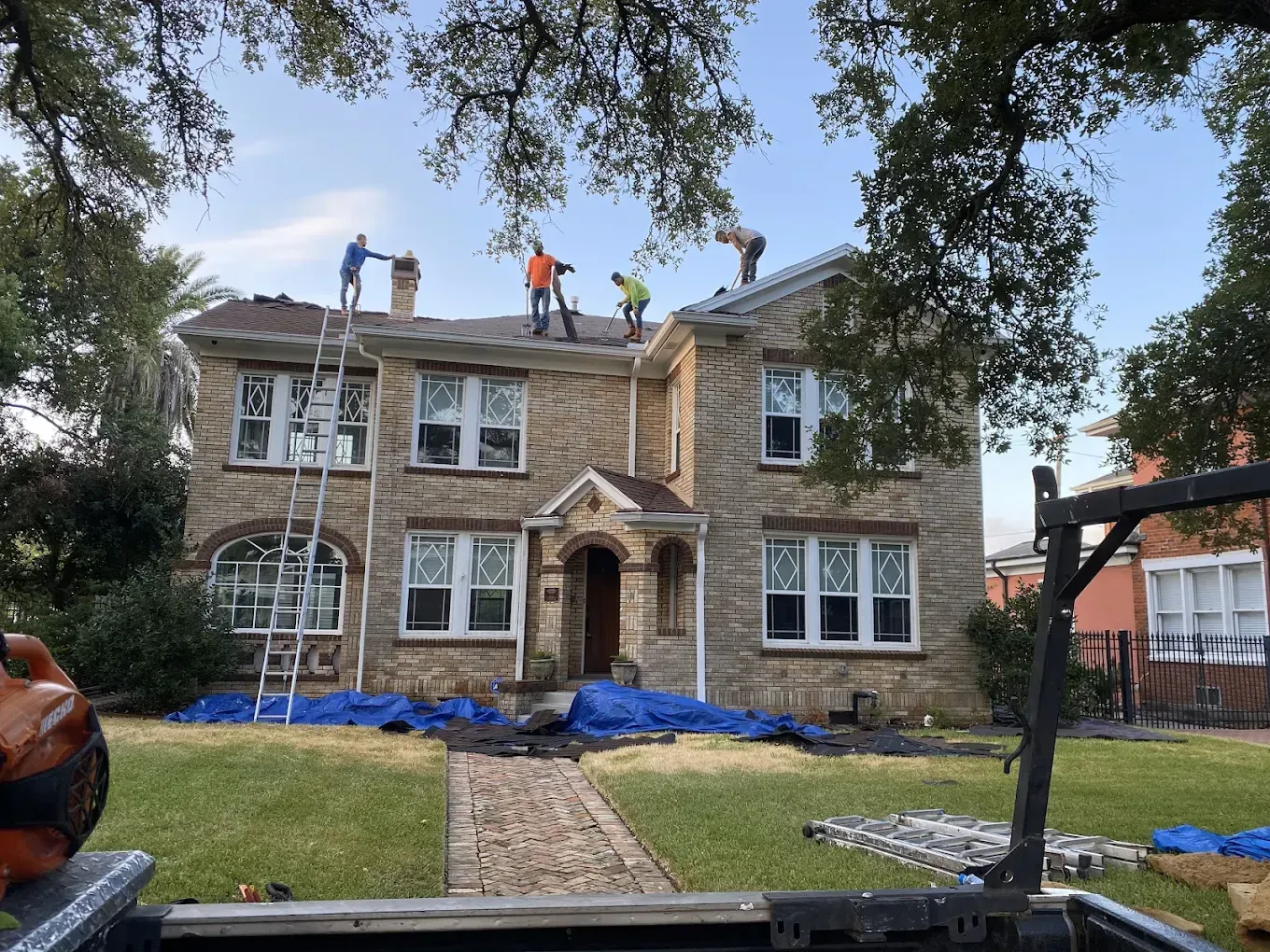Four workers replace shingles on the roof of a two-story stone house with a blue tarp spread on the lawn below.