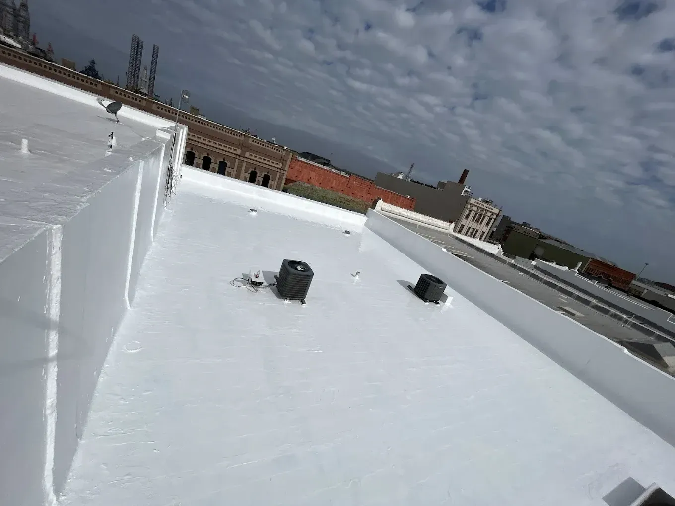 A flat, white roof surface on a building, featuring two black HVAC units and a cloudy sky in the background.