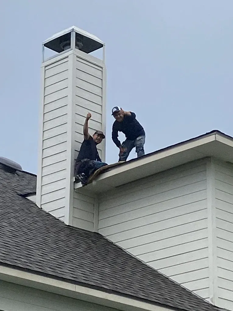 Two people on a residential roof next to a tall chimney, both waving at the camera against a clear sky.