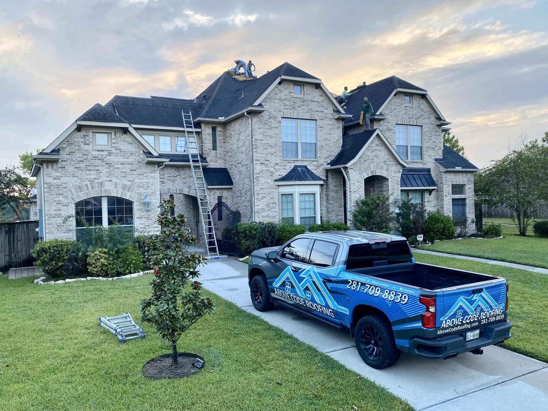A blue company truck is parked in the driveway of a two-story brick house, where workers are installing a new roof.