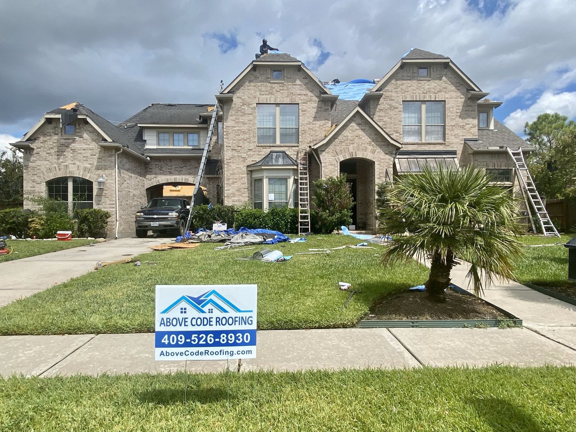 Roofers working on a multi-story brick house with a blue tarp on the roof and an Above Code Roofing sign in the yard.