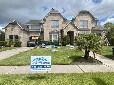 Roofers working on a multi-story brick house with a blue tarp on the roof and an Above Code Roofing sign in the yard.