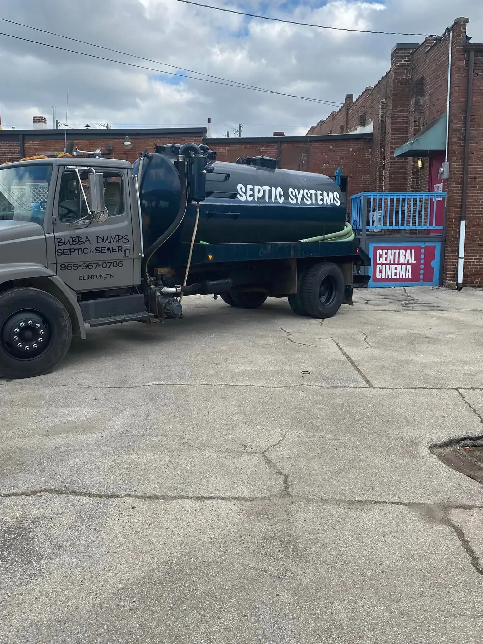 Garbage truck parked on a cracked lot beside brick buildings, with a blue dumpster and sign nearby