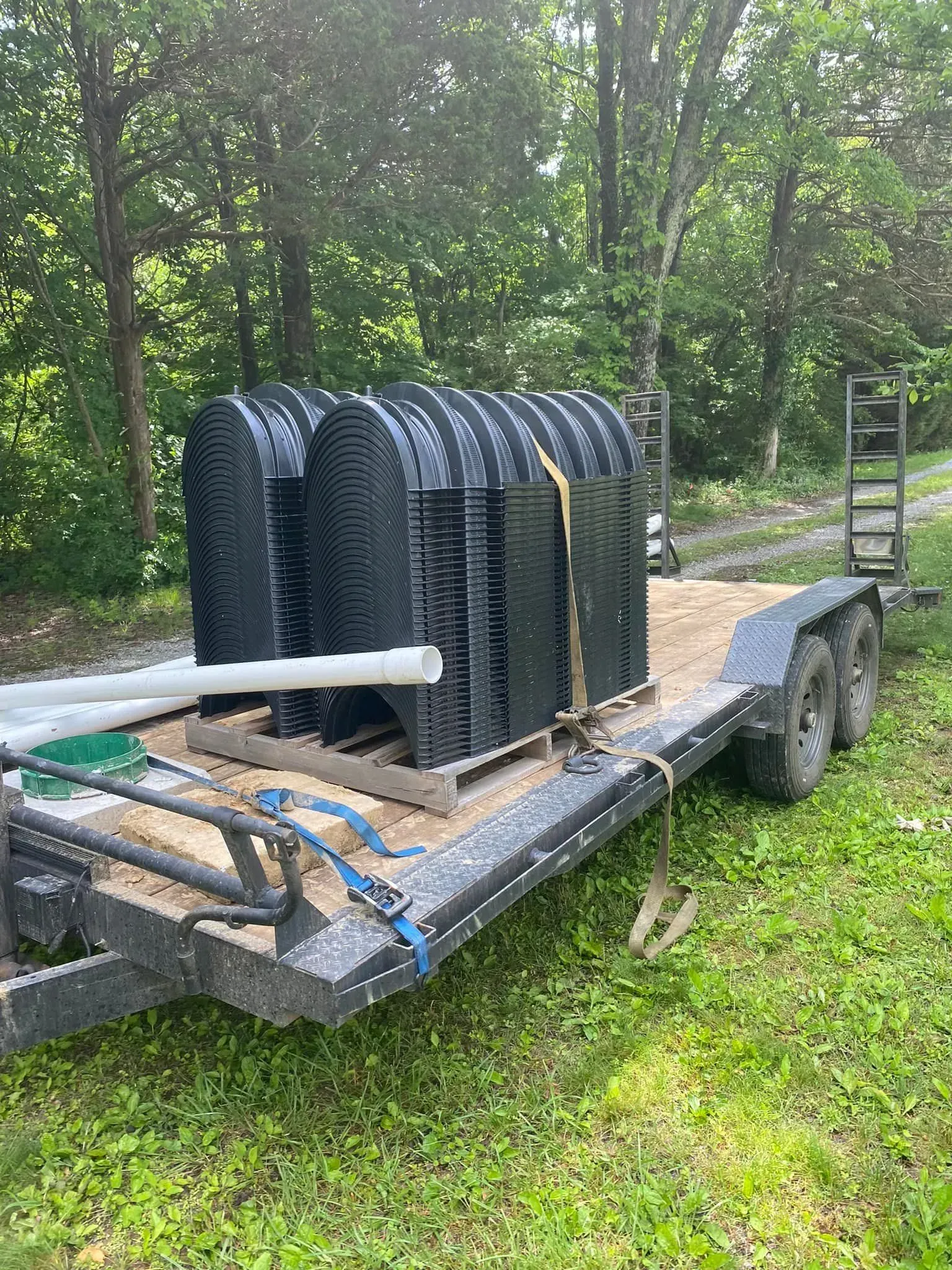 Flatbed trailer with stacked black mesh panels and a white pipe, parked on grass by trees.