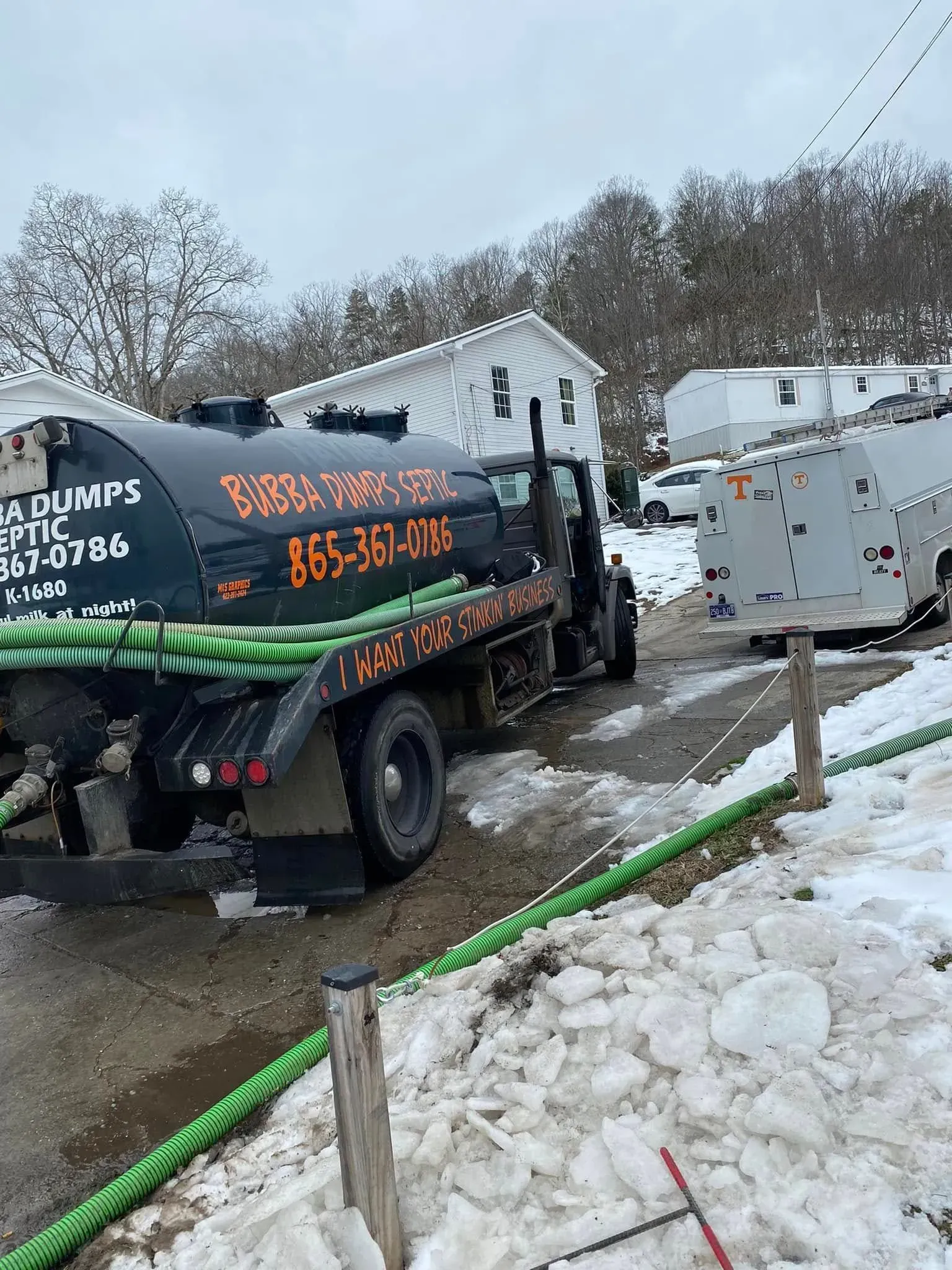 Snowy roadside with a black utility truck and a gray trailer beside a white house.