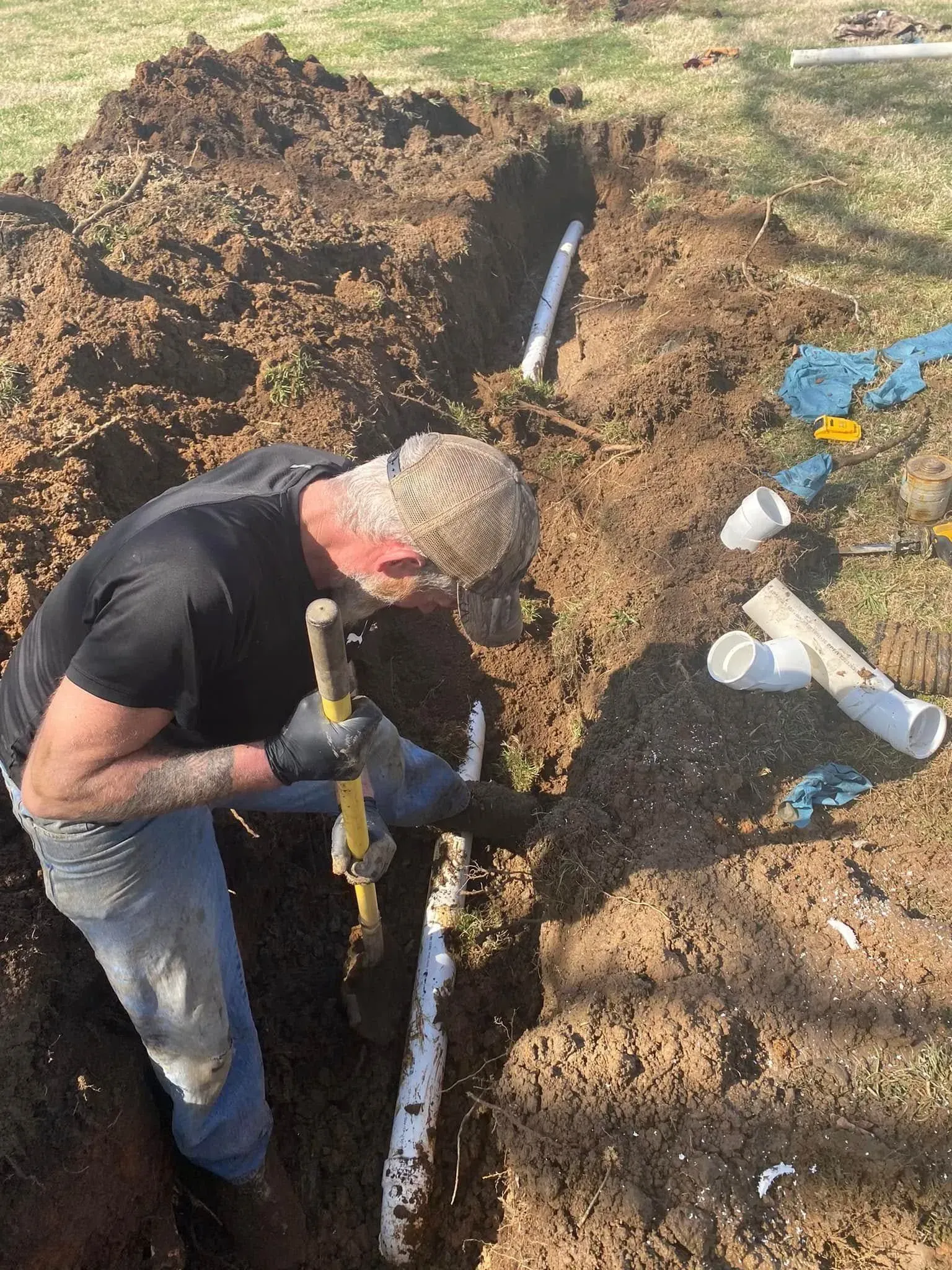 Worker digging a trench beside exposed pipes in muddy soil, with tools and water bottles nearby