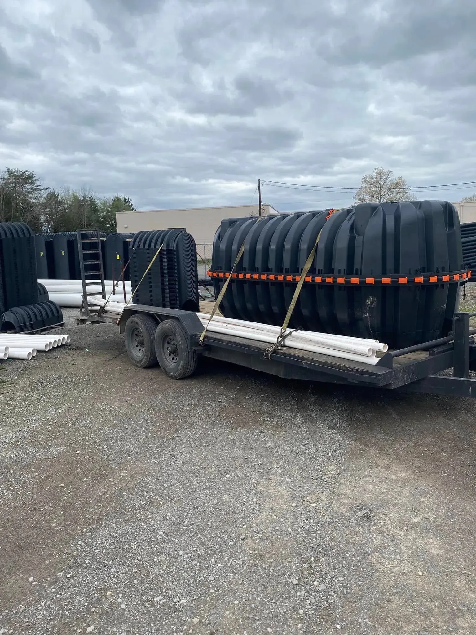 Utility trailer loaded with black coiled pipes secured by orange straps on a gravel lot under cloudy skies