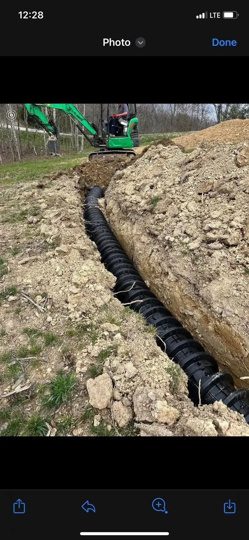 Black drainage pipe laid in a trench beside a dirt embankment, with a green excavator in the background.