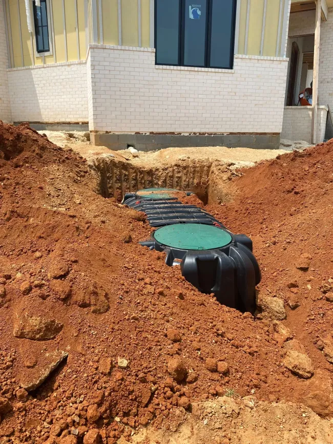 Open trench with black drainage pipes and green covers beside a building under construction.