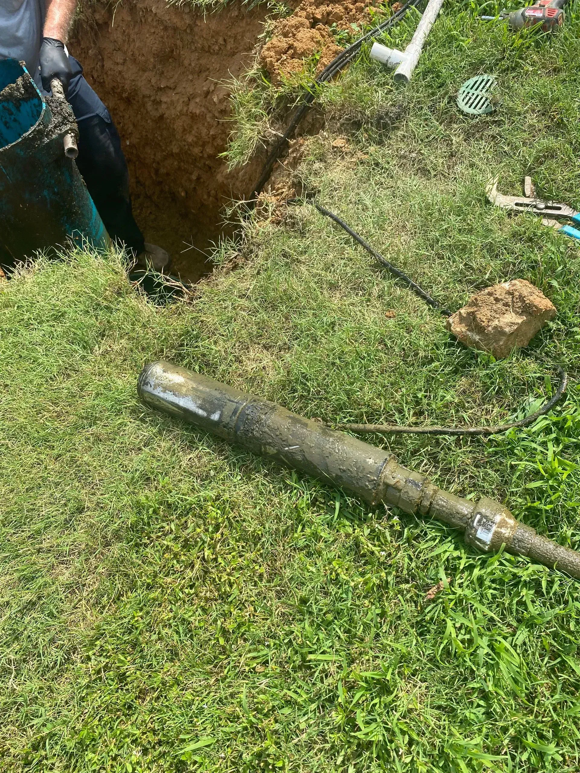 Worker beside a fresh hole in the grass, with a metal pipe and digging tools nearby