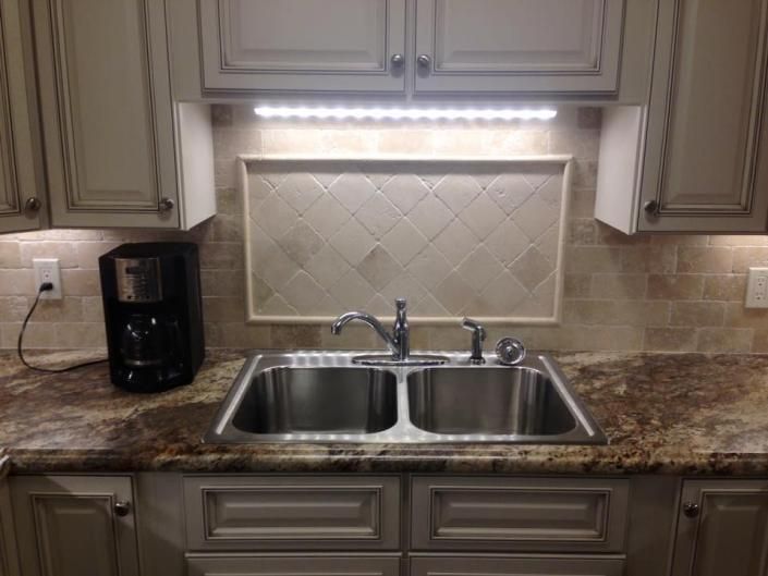 Kitchen sink with double basin, granite countertop, beige tile backsplash, and under-cabinet light.