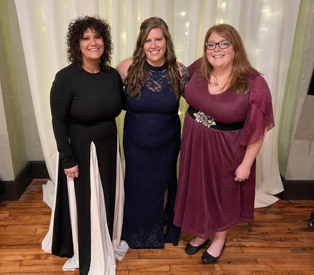 Three women standing indoors, smiling, in formal dresses on a wooden floor.