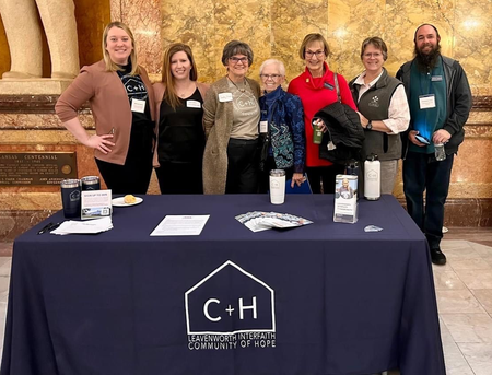 Six people standing behind a navy C+H table at a conference booth, with a gold stone wall backdrop.