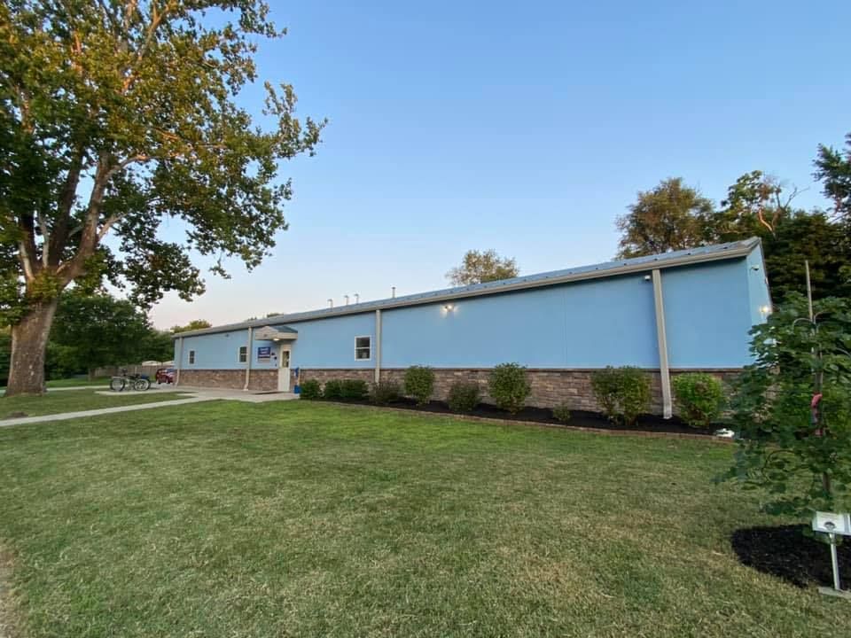 Single-story light blue building with a grassy lawn and trees under a clear sky.