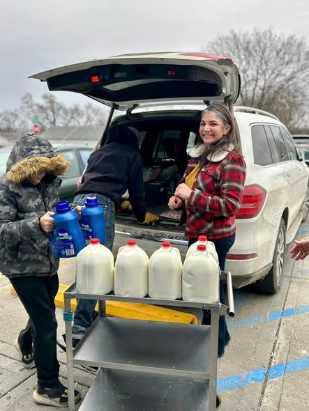 People loading jugs into SUV trunk at a curbside handout, with a smiling volunteer nearby