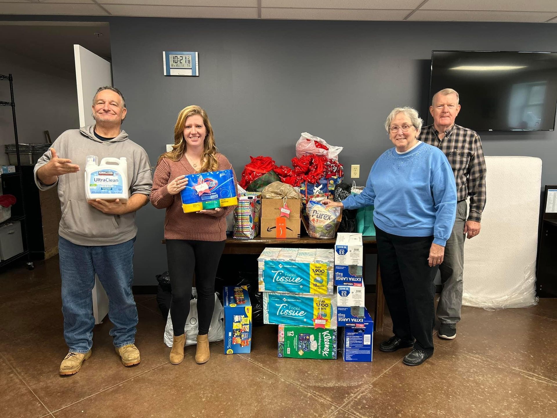 Four people standing behind a large donation pile of canned goods, snacks, and supplies indoors.