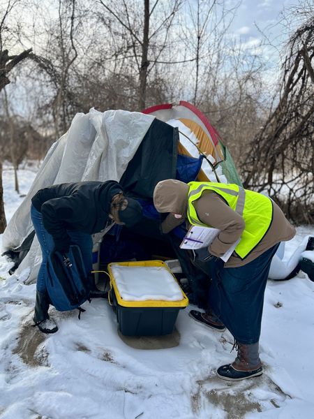 People in winter gear crouch by a tarp shelter in the snow, sorting supplies from a bin.
