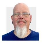 Smiling bald man with glasses and a long white goatee, wearing a blue shirt, against a white background