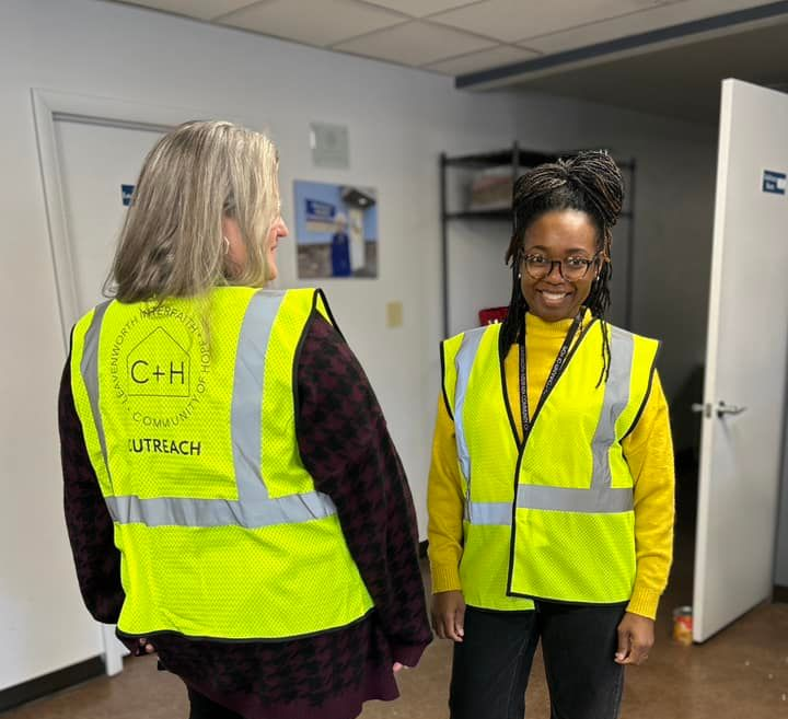 Two people in neon safety vests talking in an office hallway, one smiling at the camera.