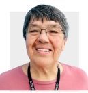 Smiling person with short gray hair wearing glasses, a black lanyard, and a pink shirt against a white background