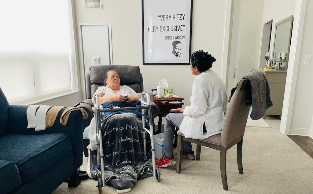 Two women sit in a room. One in a chair, one with a walker, blankets, talking. Living room setting.