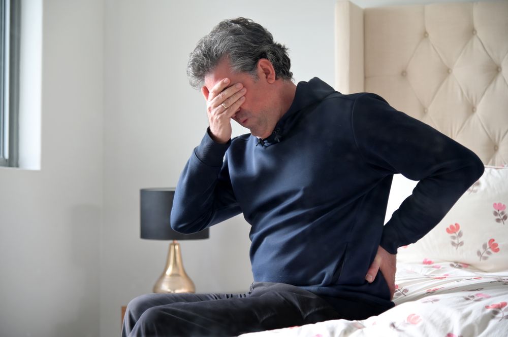 A man is sitting on a bed holding his back in pain.