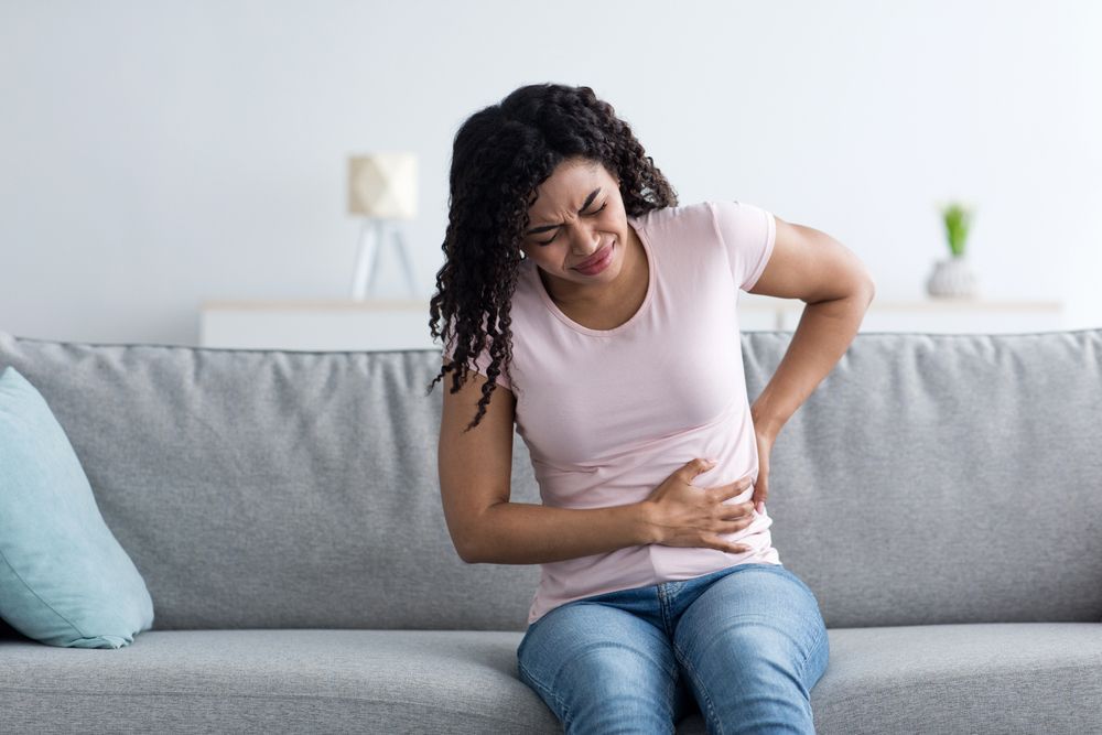 A woman is sitting on a couch holding her stomach in pain.