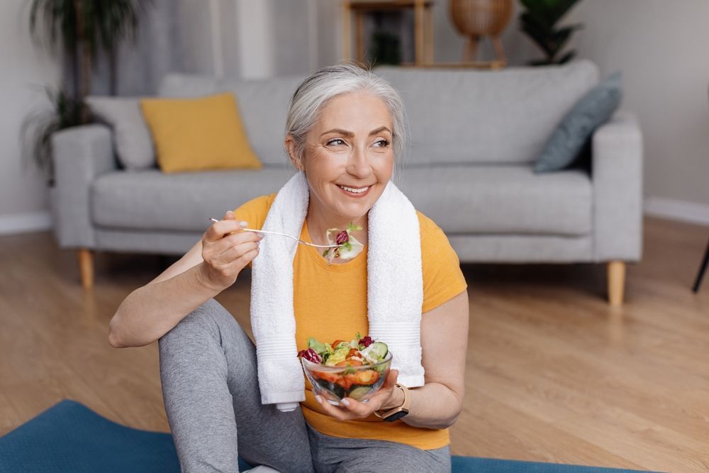 An elderly woman is sitting on a yoga mat eating a salad.