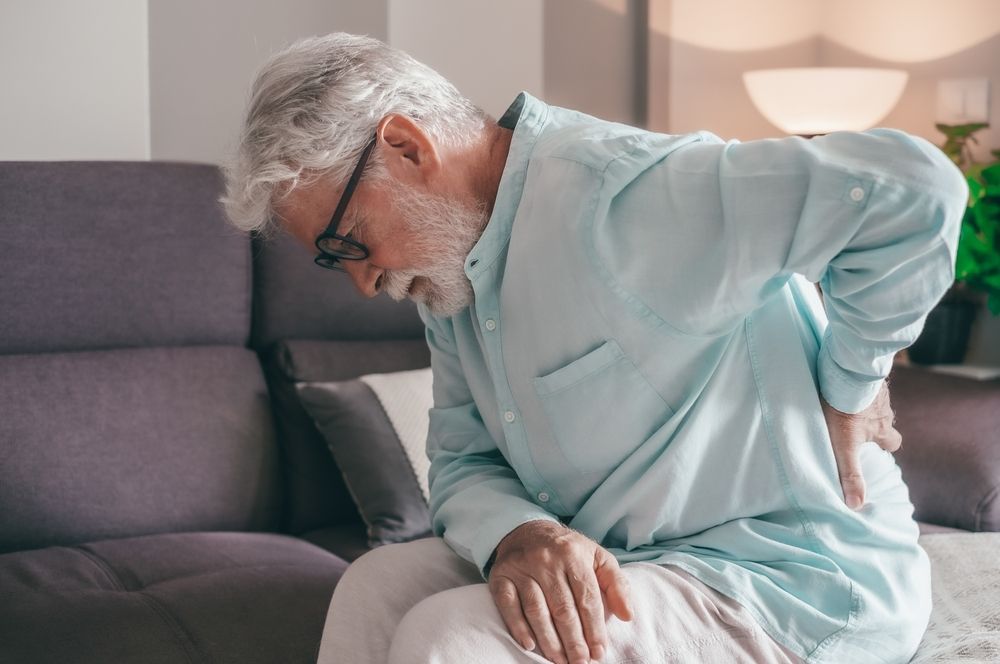 An elderly man is sitting on a couch holding his back in pain.