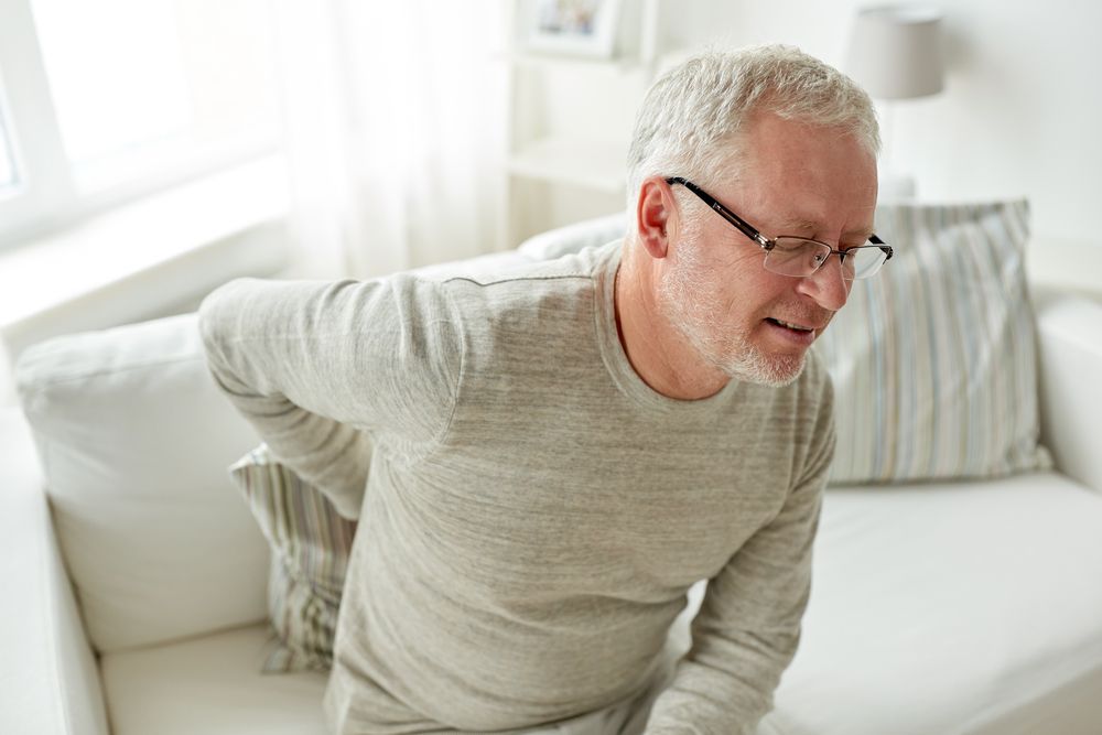 An older man is sitting on a couch holding his back in pain.