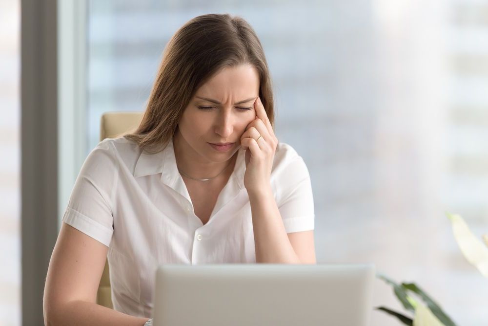 A woman is sitting in front of a laptop computer with her hand on her forehead.
