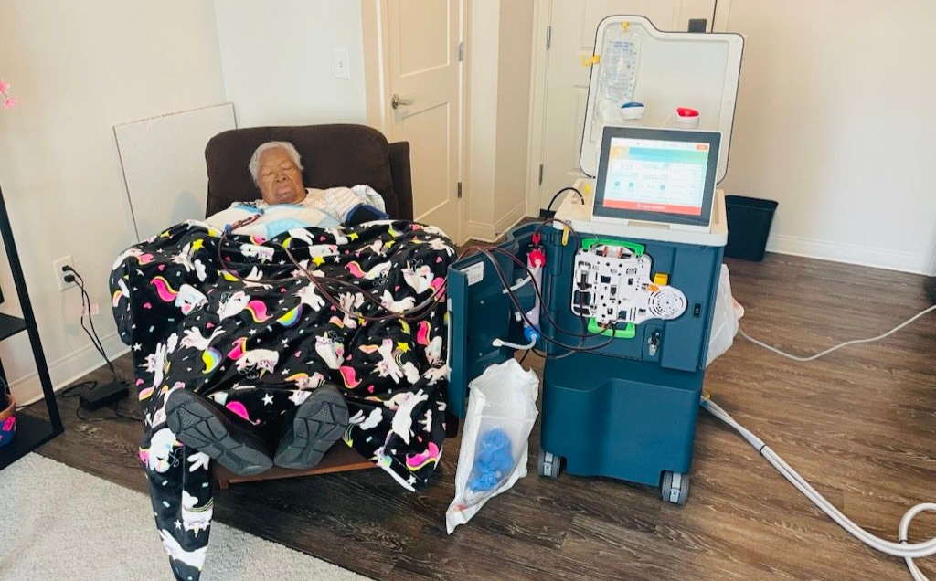 Person in chair, covered with blanket, undergoing dialysis. Machine, bag, and supplies are present.