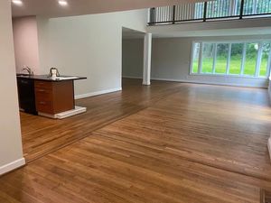 Empty open-plan room with wood floors, kitchenette, and large windows overlooking greenery