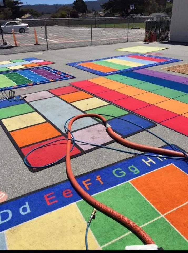 A colorful, grid-patterned educational mat lies on outdoor pavement, with a pressure washing hose snaking across it.