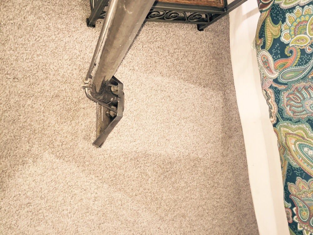 A metal carpet cleaning wand cleaning a section of beige carpet next to a patterned rug.