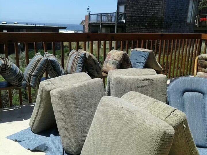 Patio cushions and throw pillows are arranged on a wooden deck to dry in the sun, with a coastal view in the background.