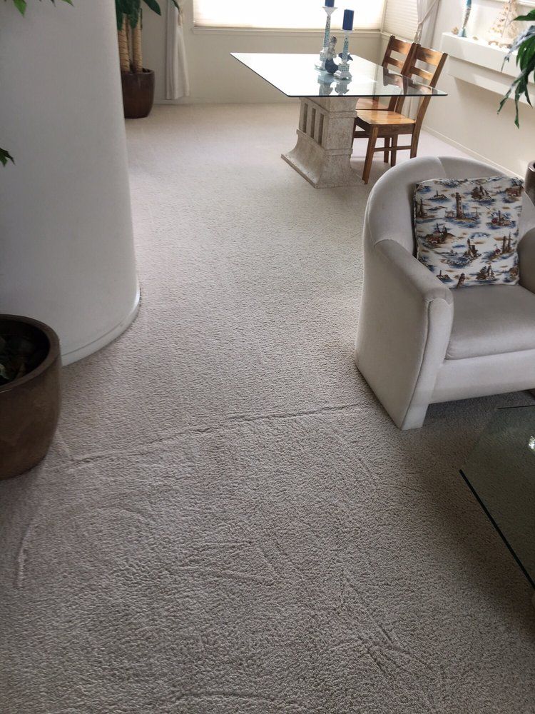 A light-colored carpeted room with an armchair, a glass dining table, and two wooden chairs.