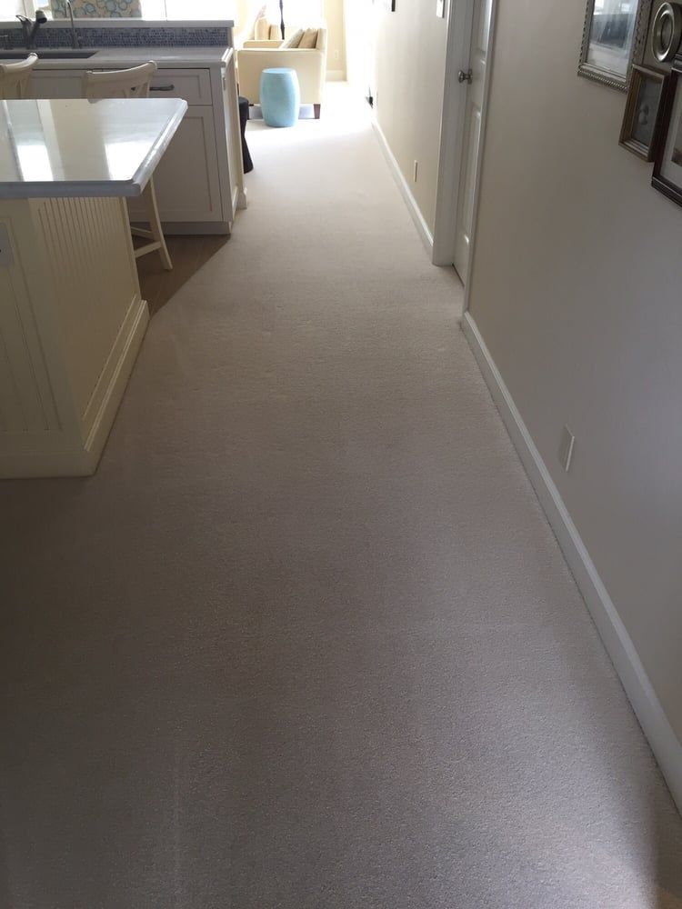 A hallway with light-colored carpet leads from a white kitchen counter toward a sunlit living area with a blue stool.