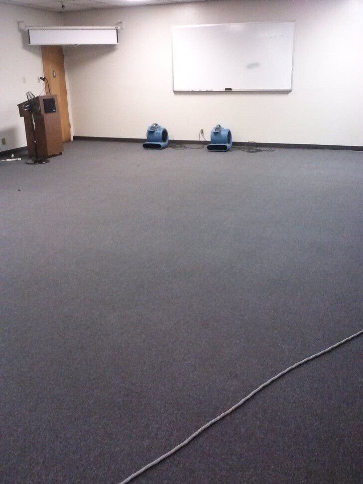An empty meeting room with gray carpet, a lectern, a whiteboard, and two blue floor drying fans.