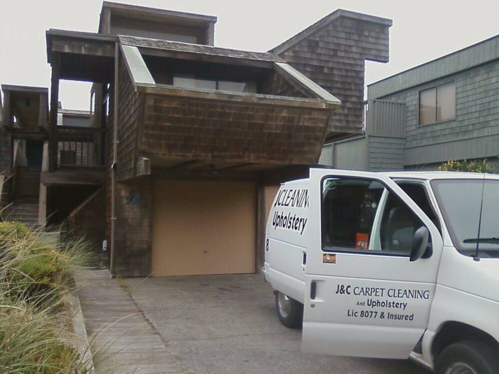 A white J&C Carpet Cleaning van parked in the driveway of a brown wooden multi-level home.