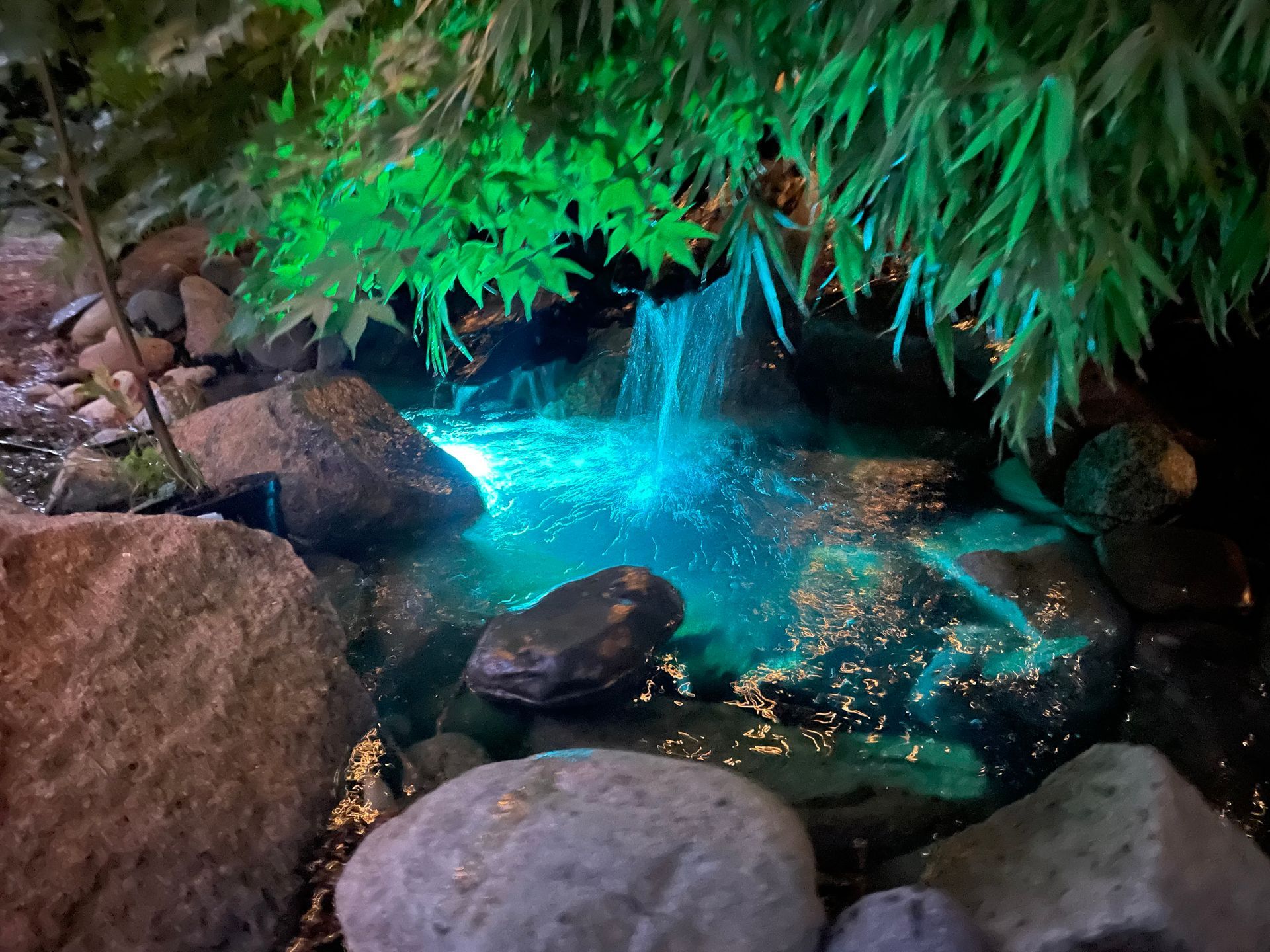 A waterfall is surrounded by rocks and trees and is lit up at night.