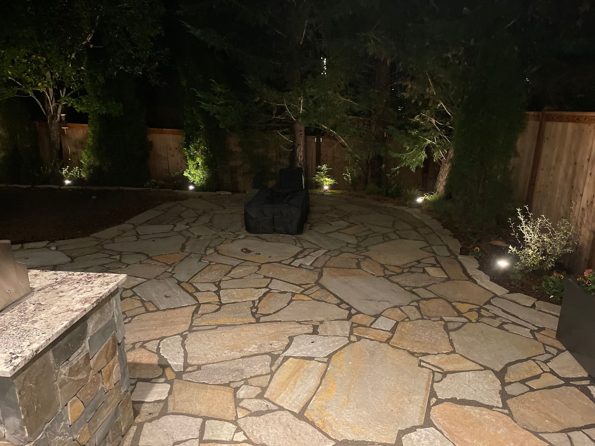 A stone patio is lit up at night with trees in the background.