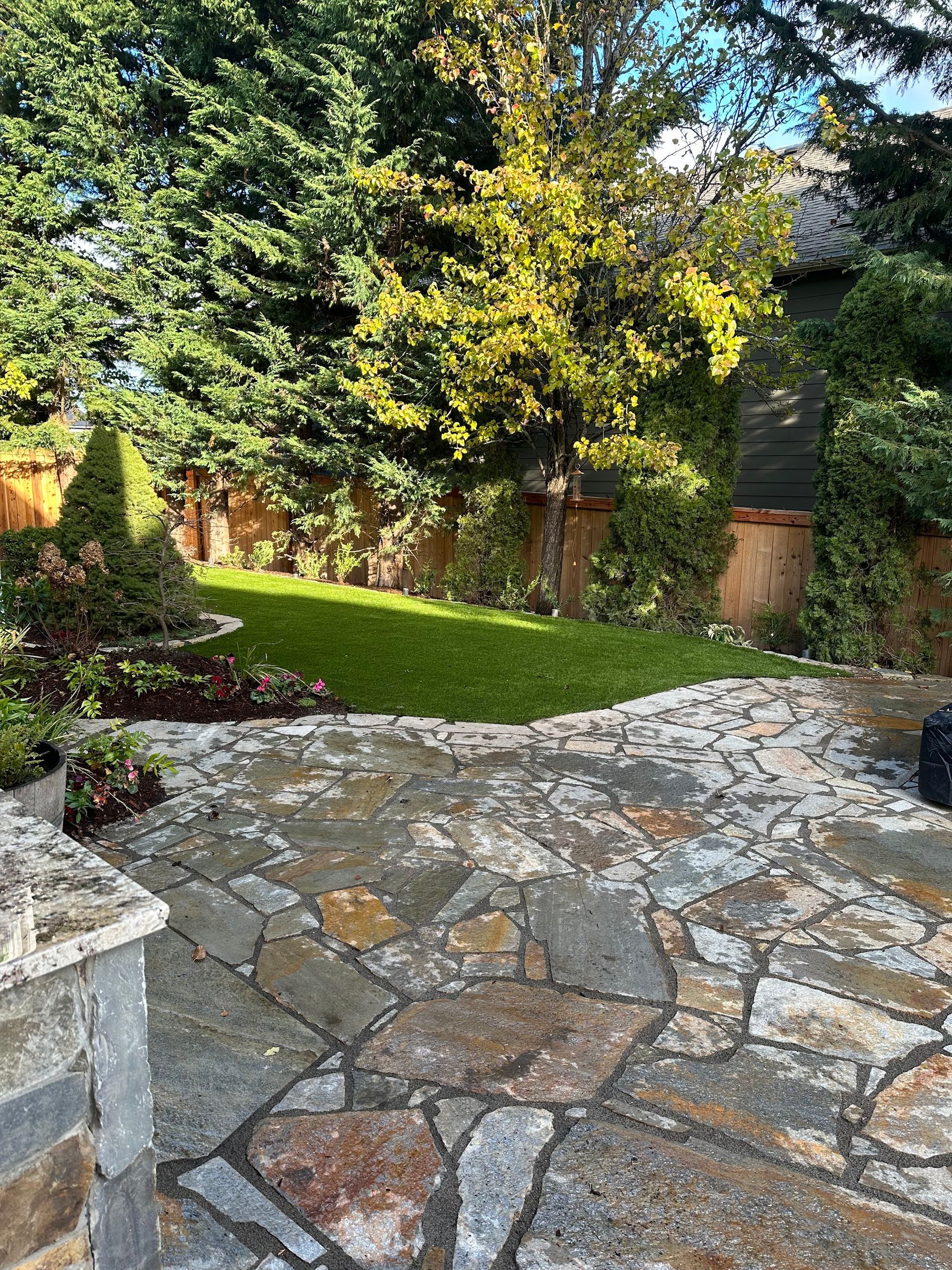 A stone patio in a backyard with a lush green lawn and trees.