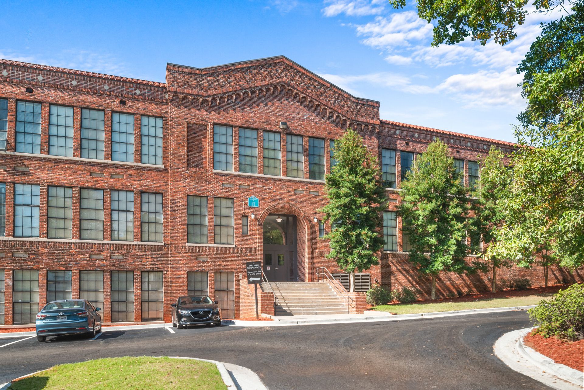 Brick apartment building with rows of windows, front steps, parked cars, and trees on a sunny day